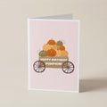 A folded greeting card standing upright on a light beige surface. The card has a pale pink background with an illustration of stacked pumpkins in a wagon and the text 'HAPPY BIRTHDAY PUMPKIN!'.