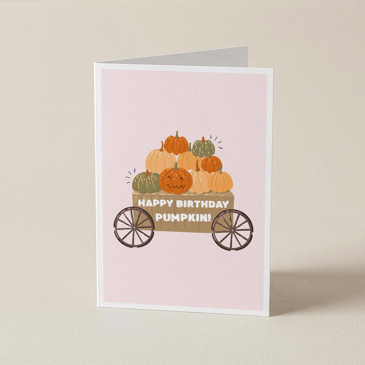 A folded greeting card standing upright on a light beige surface. The card has a pale pink background with an illustration of stacked pumpkins in a wagon and the text 'HAPPY BIRTHDAY PUMPKIN!'.