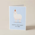 A folded greeting card standing upright on a light beige surface. The card features a white sheep wearing a small orange party hat on a pale blue background, with the text 'HAPPY BIRTHDAY TO EWE' printed below.