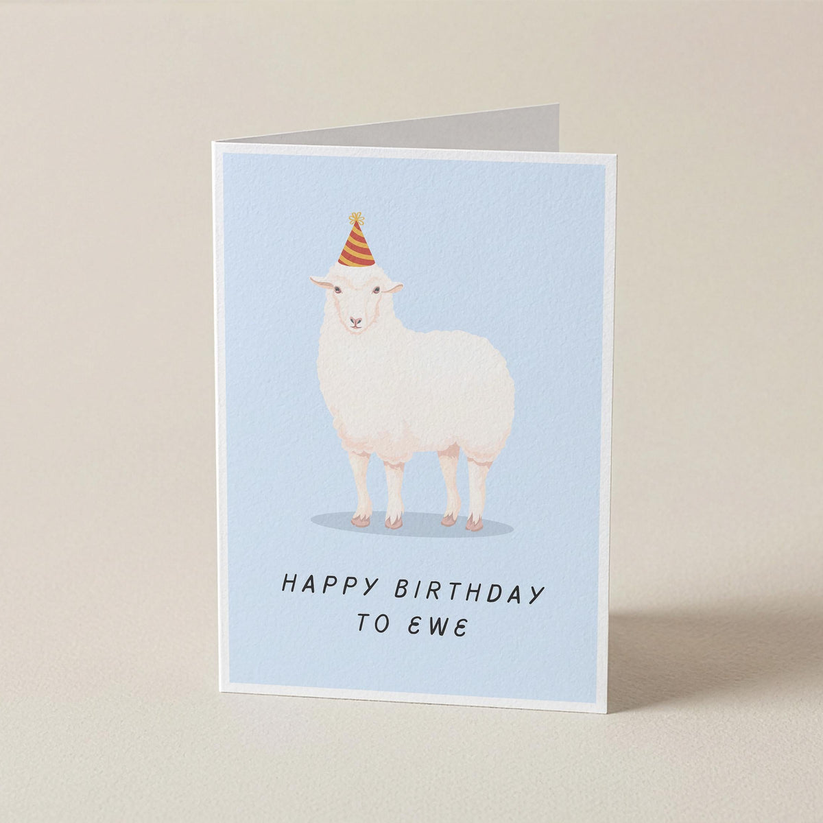 A folded greeting card standing upright on a light beige surface. The card features a white sheep wearing a small orange party hat on a pale blue background, with the text 'HAPPY BIRTHDAY TO EWE' printed below.