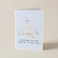 A folded greeting card standing upright on a light beige surface. The card features a pale blue front with a minimalist illustration of a white goose with a blue bow, two small yellow chicks, and the text 'WELCOME TO THE WORLD, LITTLE ONE.