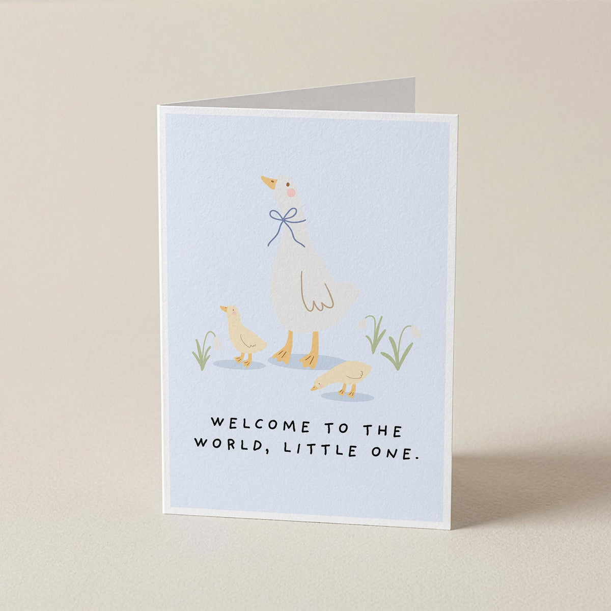 A folded greeting card standing upright on a light beige surface. The card features a pale blue front with a minimalist illustration of a white goose with a blue bow, two small yellow chicks, and the text 'WELCOME TO THE WORLD, LITTLE ONE.