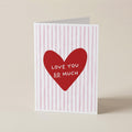 A folded greeting card standing upright on a light beige surface. The card has a white background with vertical pink stripes and a large red heart in the center containing the white text 'LOVE YOU SO MUCH'.