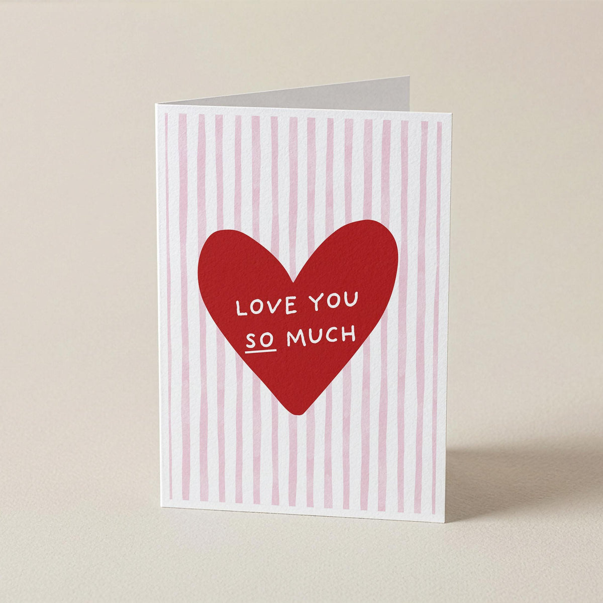 A folded greeting card standing upright on a light beige surface. The card has a white background with vertical pink stripes and a large red heart in the center containing the white text 'LOVE YOU SO MUCH'.