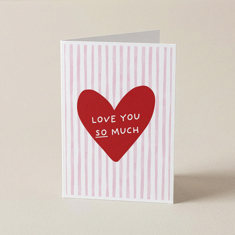 A folded greeting card standing upright on a light beige surface. The card has a white background with vertical pink stripes and a large red heart in the center containing the white text 'LOVE YOU SO MUCH'.