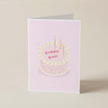 A folded greeting card standing upright on a light beige surface. The card has a pale pink background with an illustration of a birthday cake topped with two lit candles and the words 'Birthday Queen' in pink lettering.