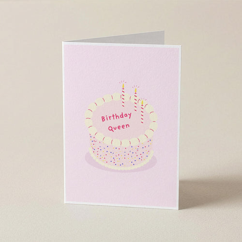 A folded greeting card standing upright on a light beige surface. The card has a pale pink background with an illustration of a birthday cake topped with two lit candles and the words 'Birthday Queen' in pink lettering.
