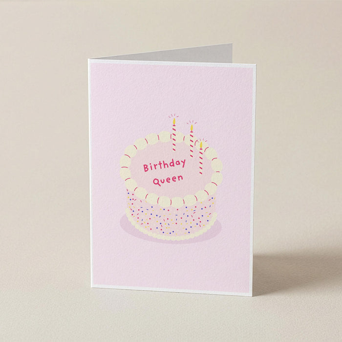 A folded greeting card standing upright on a light beige surface. The card has a pale pink background with an illustration of a birthday cake topped with two lit candles and the words 'Birthday Queen' in pink lettering.