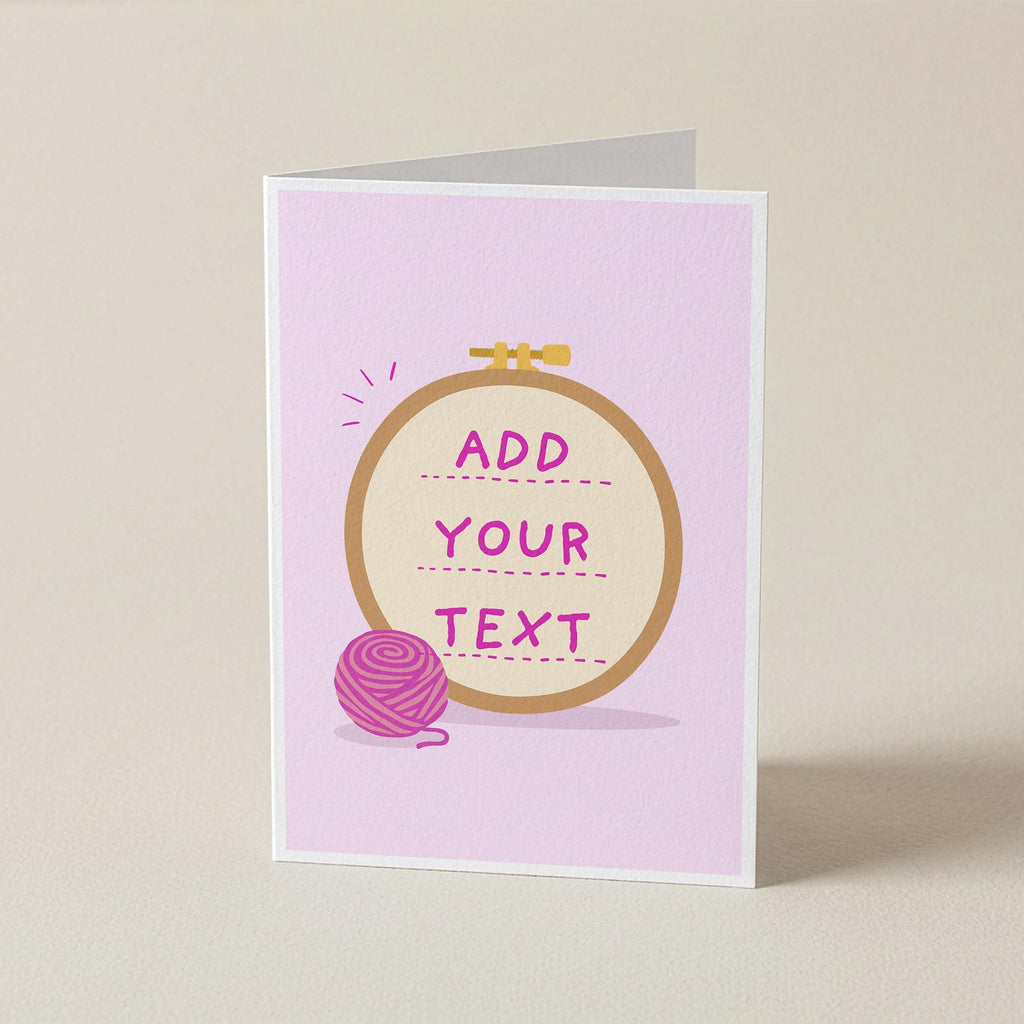 A folded greeting card standing upright on a light beige surface. The card has a pale pink background with a central circular embroidery hoop illustration in tan and gold, pink text reading 'ADD YOUR TEXT,' and a pink yarn ball graphic.
