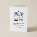 A white square congratulations card standing upright on a light beige surface. The card features a simple black line drawing of two raised hands with small gold rings, a small red heart, and the text 'CONGRATULATIONS CHARLES & RACHEL! 05.