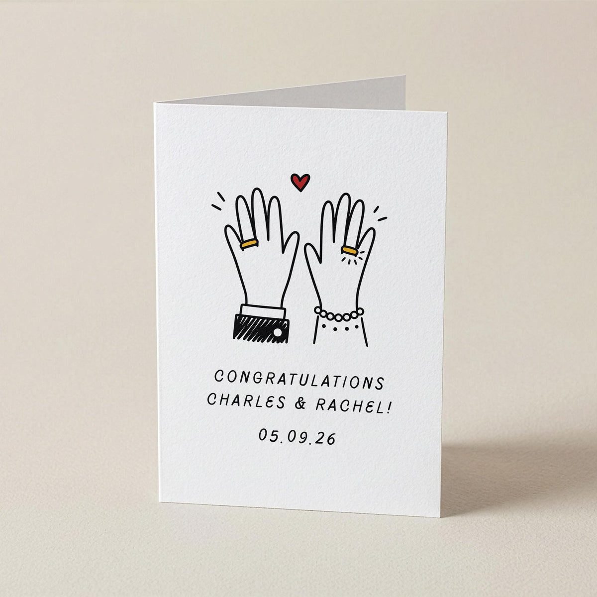 A white square congratulations card standing upright on a light beige surface. The card features a simple black line drawing of two raised hands with small gold rings, a small red heart, and the text 'CONGRATULATIONS CHARLES & RACHEL! 05.