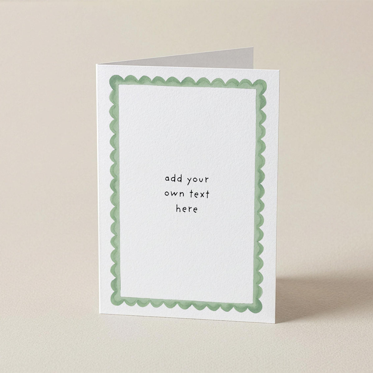A white greeting card with a light green scalloped border and placeholder text reading 'add your own text here', standing upright on a beige background.