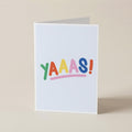 A white greeting card standing upright on a beige surface, printed with the word 'YAAAS!' in colorful playful letters and an exclamation mark.