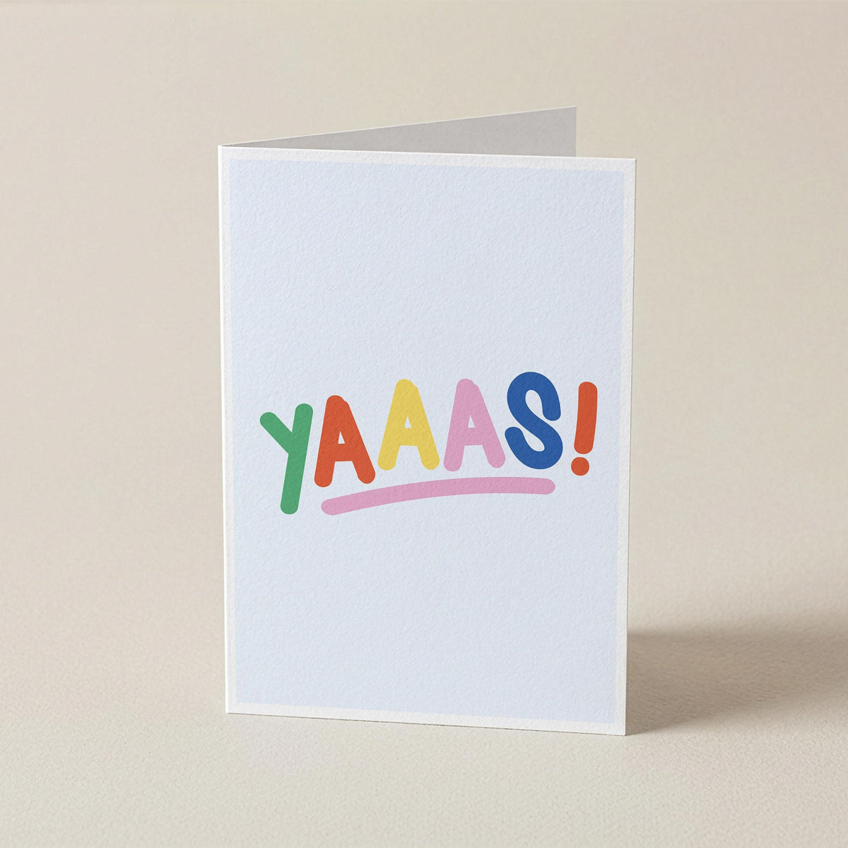 A white greeting card standing upright on a beige surface, printed with the word 'YAAAS!' in colorful playful letters and an exclamation mark.