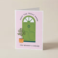 A folded greeting card standing upright on a light beige surface. The card has a pale pink background with an illustration of a green front door, a small potted plant, and black text reading 'SHUT THE FRONT DOOR!' and 'YOU BOUGHT A HOUSE!'.