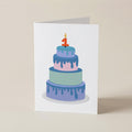 A white greeting card standing upright on a beige surface, printed with an illustration of a three-tier cake in pastel blue, purple, and mint with a red number 1 candle.