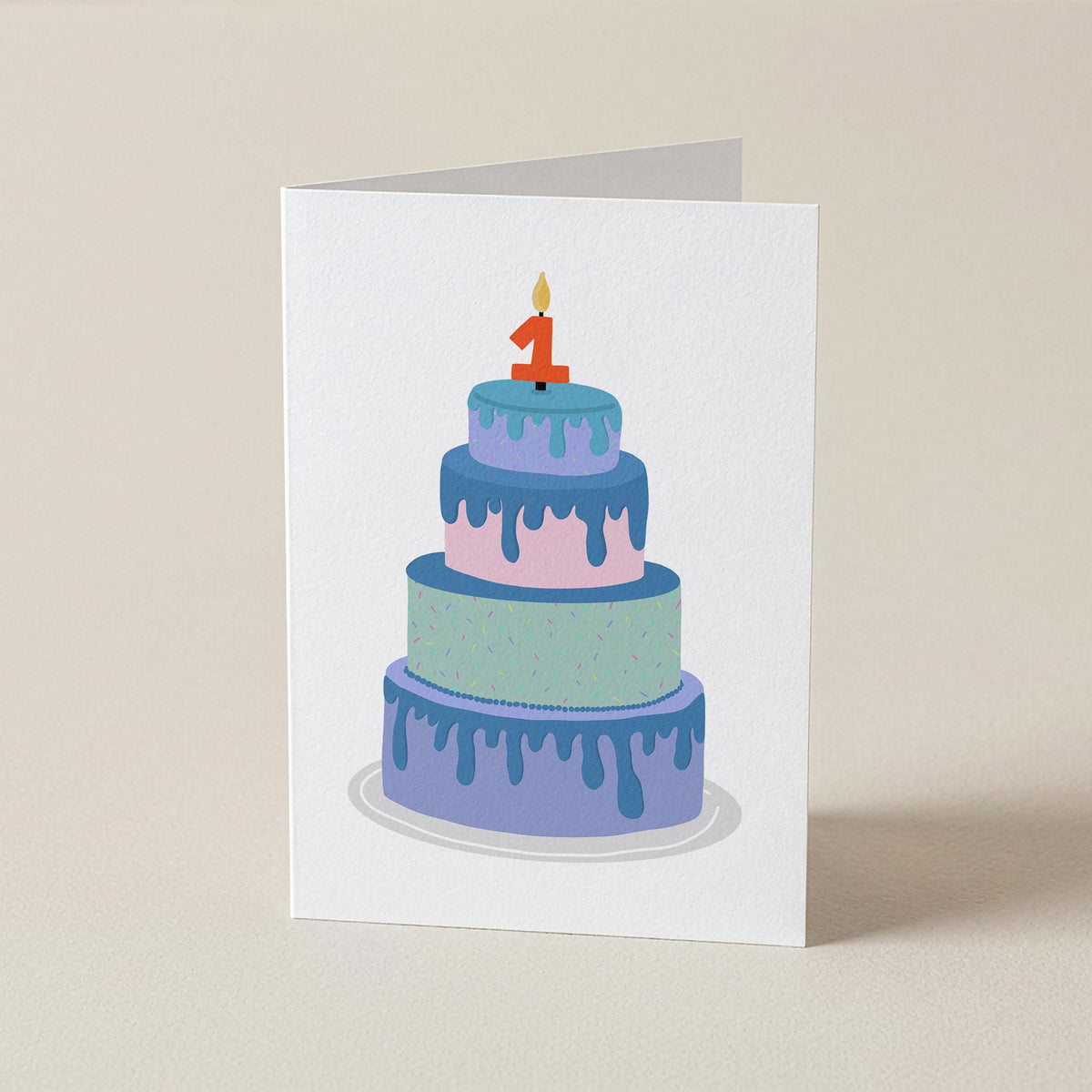 A white greeting card standing upright on a beige surface, printed with an illustration of a three-tier cake in pastel blue, purple, and mint with a red number 1 candle.