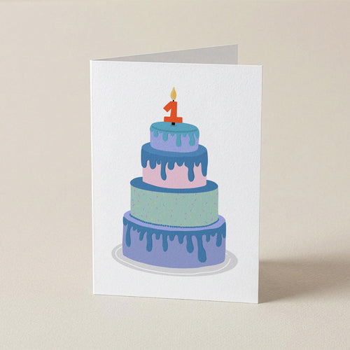 A white greeting card standing upright on a beige surface, printed with an illustration of a three-tier cake in pastel blue, purple, and mint with a red number 1 candle.