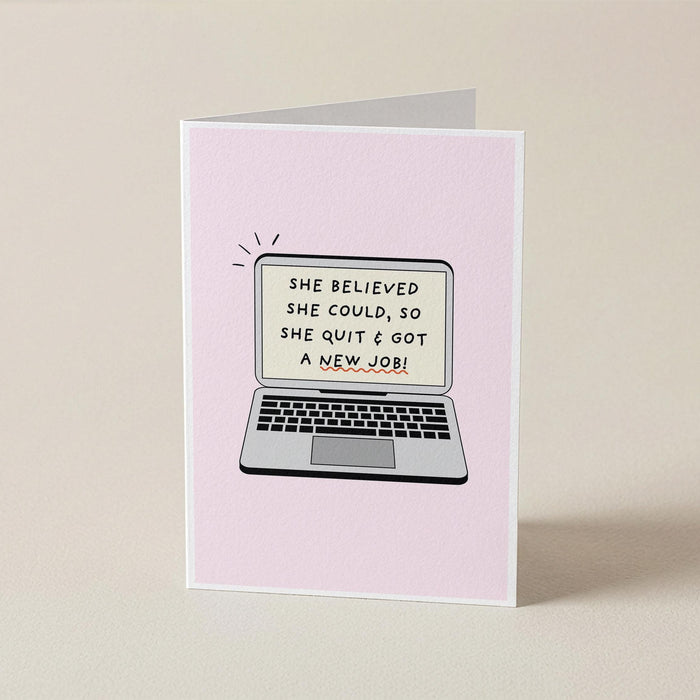 A folded greeting card standing upright on a light beige surface. The card front is pale pink and features an illustration of a laptop with the text, 'SHE BELIEVED SHE COULD, SO SHE QUIT & GOT A NEW JOB!'.