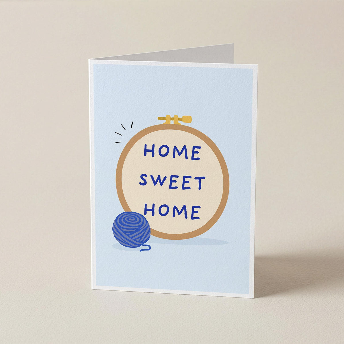 A folded greeting card standing upright on a light beige surface. The card has a pale blue background and features a central circular embroidery-style design with the words “HOME SWEET HOME” in blue lettering inside a tan circle, plus a small blue ball of yarn and a tiny gold bow detail.
