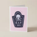 A folded greeting card standing upright on a light beige surface. The card has a pink front with a dark navy tombstone graphic featuring a white skull and crossbones and the text 'R.I.P 20s'.