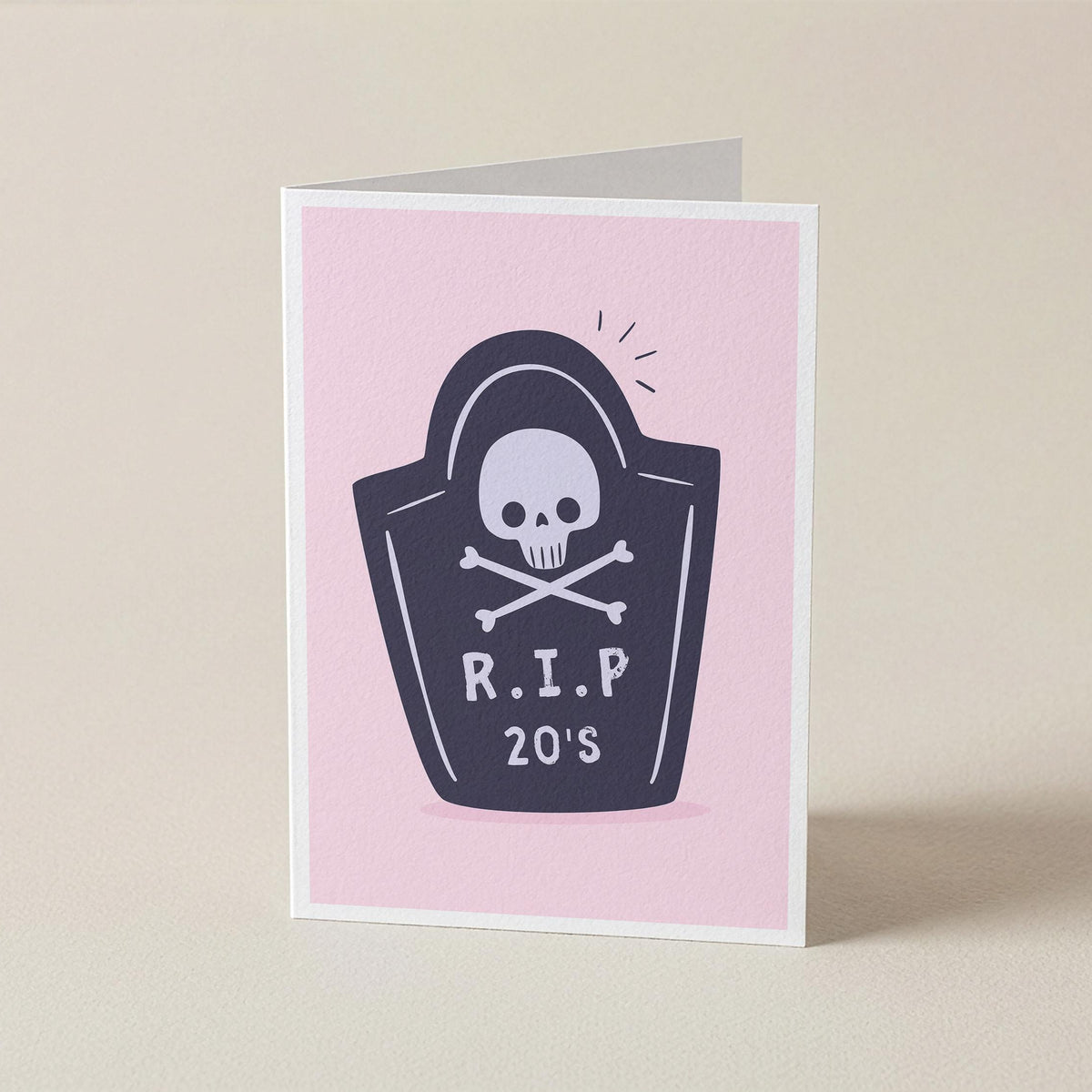 A folded greeting card standing upright on a light beige surface. The card has a pink front with a dark navy tombstone graphic featuring a white skull and crossbones and the text 'R.I.P 20s'.