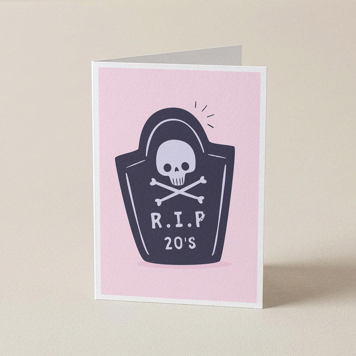 A folded greeting card standing upright on a light beige surface. The card has a pink front with a dark navy tombstone graphic featuring a white skull and crossbones and the text 'R.I.P 20s'.