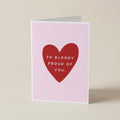 A folded greeting card standing upright on a light beige surface. The card has a pale pink background with a large red heart in the center containing the white text 'SO BLOODY PROUD OF YOU.