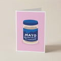 A folded greeting card standing upright on a light beige surface. The card features a pink background with a stylized illustration of a jar labeled 'MAYO BIRTHDAY BE FULL OF HAPPINESS'.