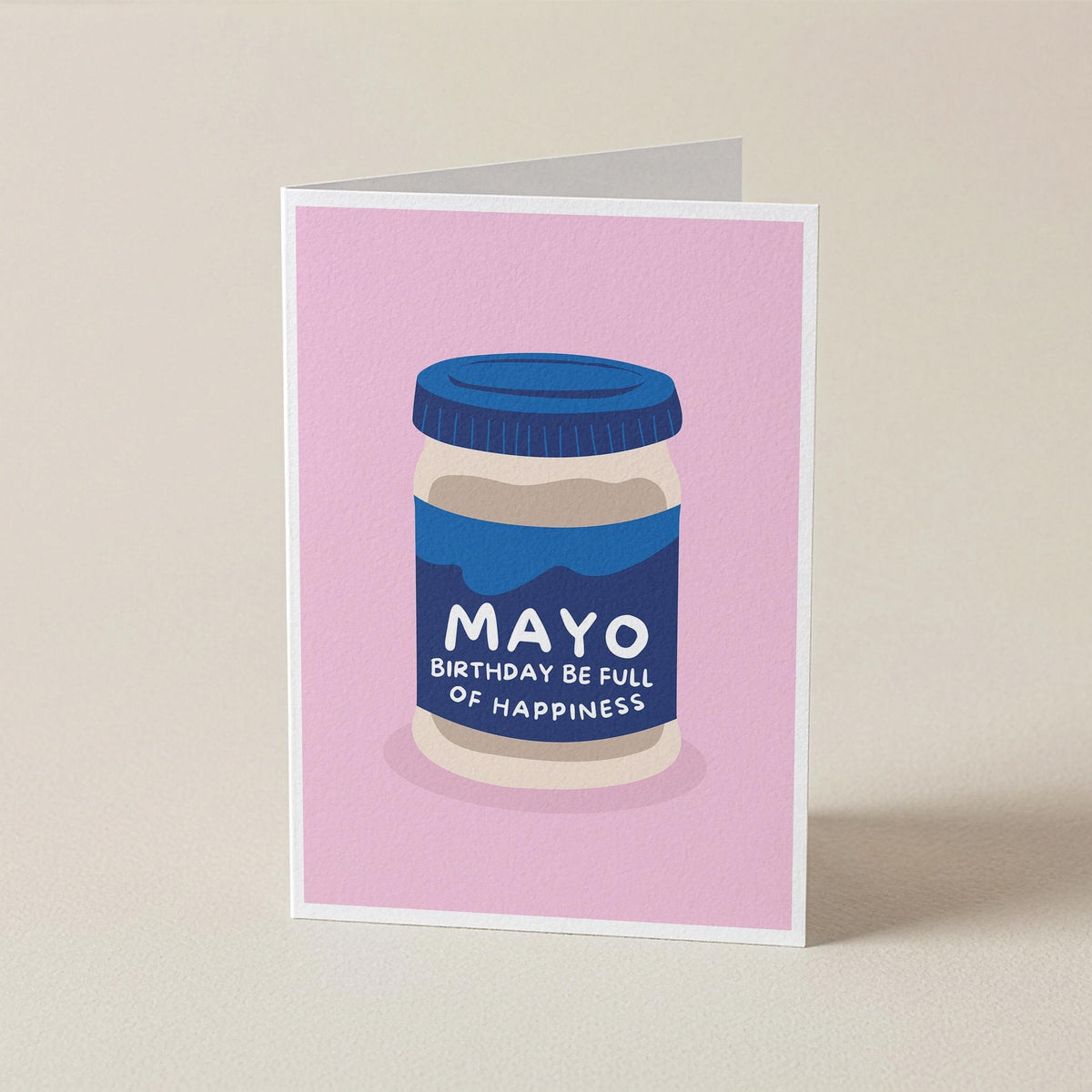 A folded greeting card standing upright on a light beige surface. The card features a pink background with a stylized illustration of a jar labeled 'MAYO BIRTHDAY BE FULL OF HAPPINESS'.