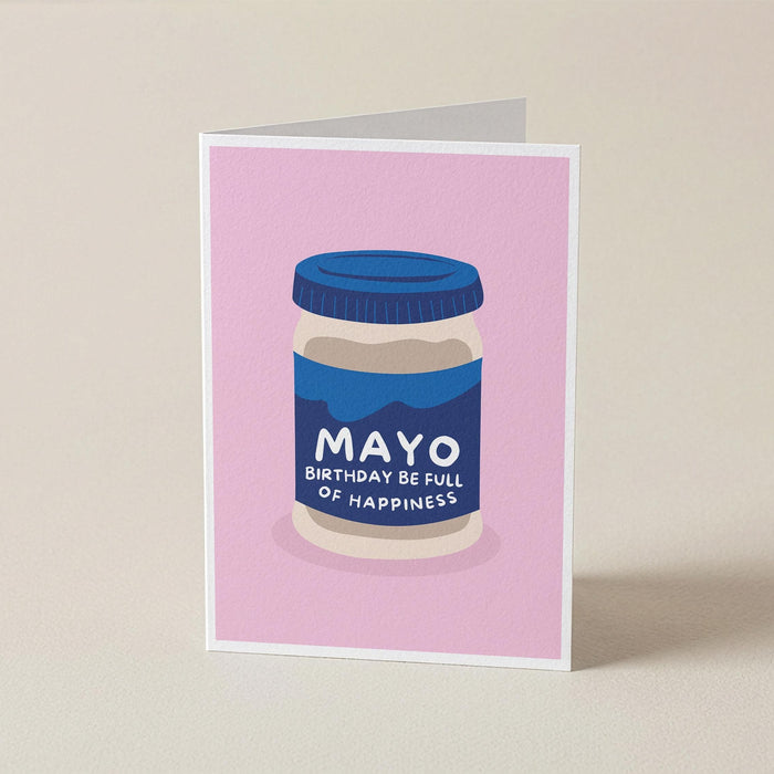 A folded greeting card standing upright on a light beige surface. The card features a pink background with a stylized illustration of a jar labeled 'MAYO BIRTHDAY BE FULL OF HAPPINESS'.