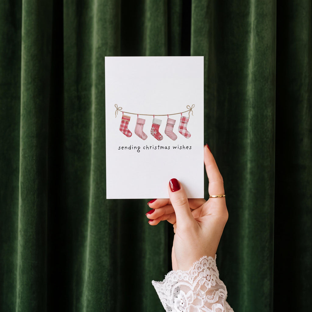 A hand with red nail polish and a white lace sleeve holds a white greeting card featuring a watercolor illustration of red and pink Christmas stockings on a string, with the text 'sending christmas wishes' below. The background is a dark green velvet curtain.