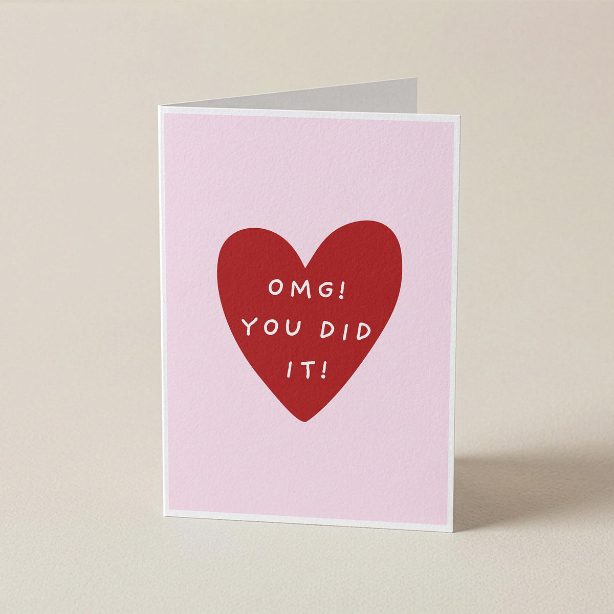 A folded greeting card standing upright on a light beige surface. The card has a pale pink background with a large red heart in the center and the words 'Omg! You did it!'