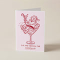 A folded greeting card standing upright on a light beige surface. The card is pale pink with a red line drawing of Santa Claus sitting in a martini glass and holding a candy cane, with the text 'TIS THE SEASON FOR COCKTAILS!' printed below.