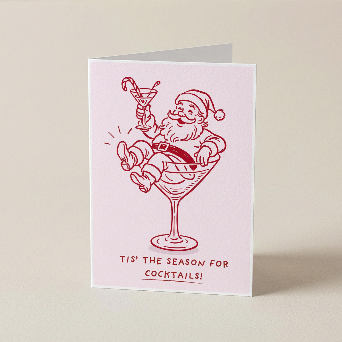 A folded greeting card standing upright on a light beige surface. The card is pale pink with a red line drawing of Santa Claus sitting in a martini glass and holding a candy cane, with the text 'TIS THE SEASON FOR COCKTAILS!' printed below.