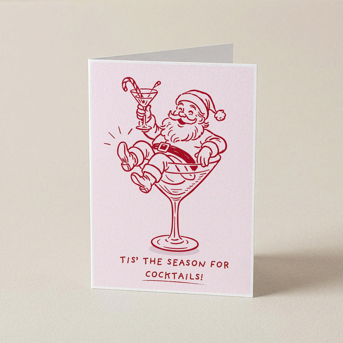 A folded greeting card standing upright on a light beige surface. The card is pale pink with a red line drawing of Santa Claus sitting in a martini glass and holding a candy cane, with the text 'TIS THE SEASON FOR COCKTAILS!' printed below.