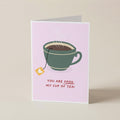 A folded greeting card standing upright on a light beige surface. The card has a pale pink background with an illustration of a green teacup with a tea bag and the text 'YOU ARE SOON MY CUP OF TEA!'.