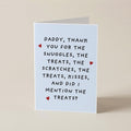 A folded greeting card standing upright on a plain beige surface. The card is white with black handwritten-style text reading, 'Daddy, thank you for the snuggles, the treats, the scratches, the treats, kisses, and did I mention the treats?' and small red heart accents.