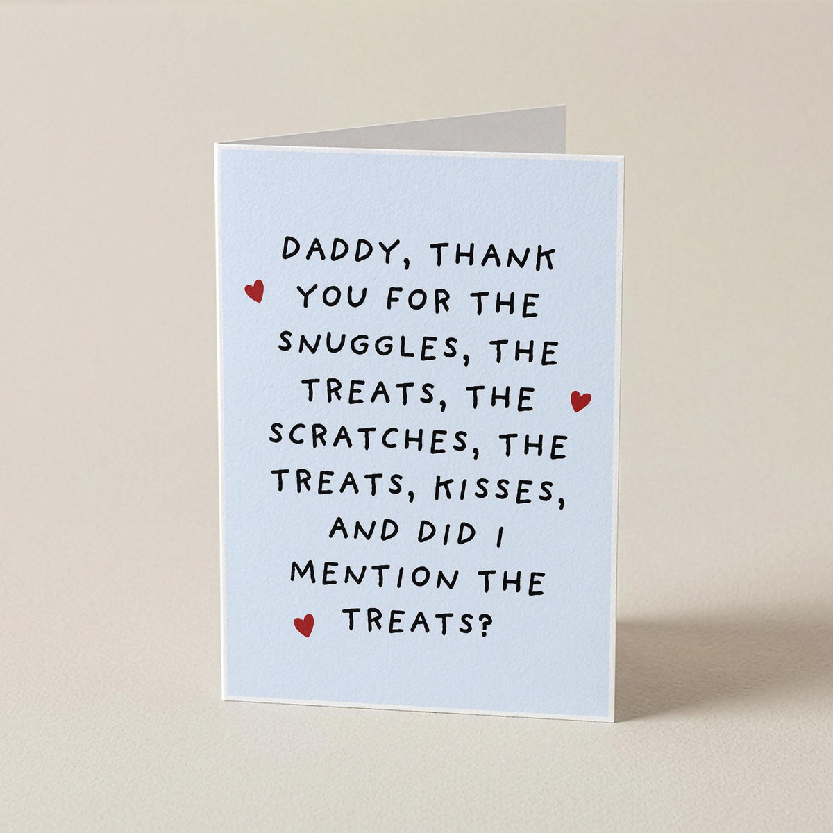 A folded greeting card standing upright on a plain beige surface. The card is white with black handwritten-style text reading, 'Daddy, thank you for the snuggles, the treats, the scratches, the treats, kisses, and did I mention the treats?' and small red heart accents.