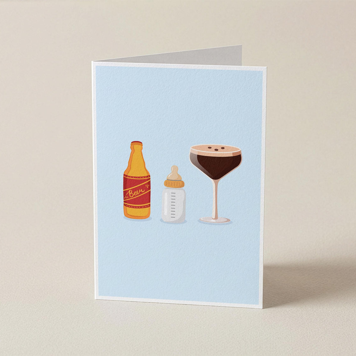 A folded greeting card standing upright on a beige surface, featuring a light blue background with illustrated baby bottle and cocktail glass icons.