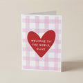 A folded greeting card standing upright on a light beige surface. The card has a white and pale pink gingham pattern, with a large red heart in the center containing the white text 'WELCOME TO THE WORLD, OLIVE'.