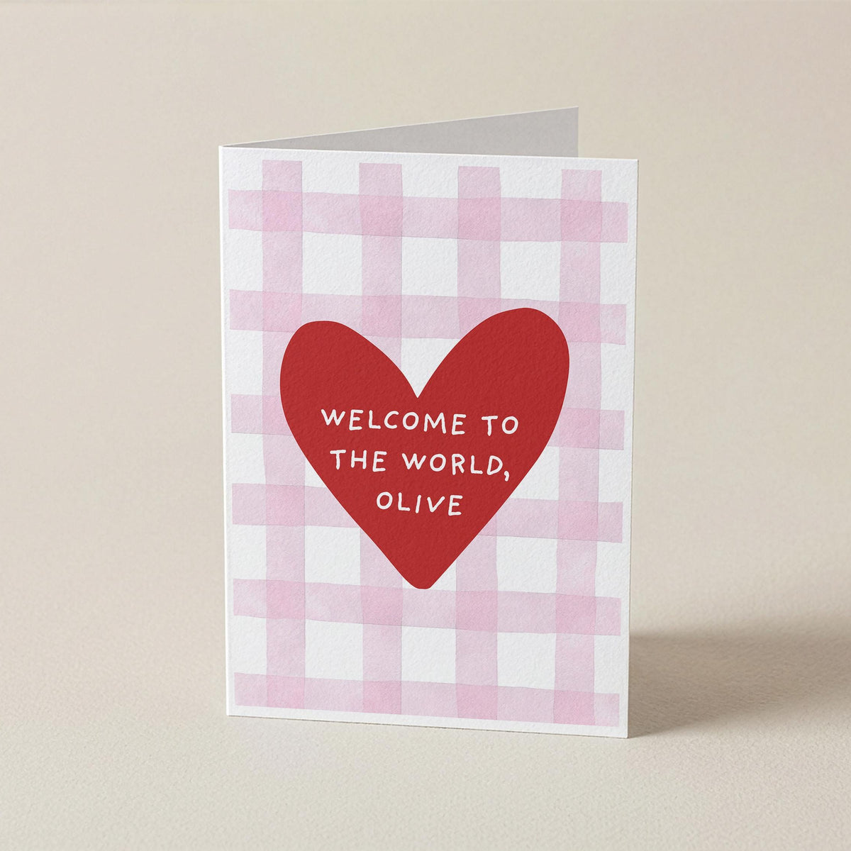 A folded greeting card standing upright on a light beige surface. The card has a white and pale pink gingham pattern, with a large red heart in the center containing the white text 'WELCOME TO THE WORLD, OLIVE'.