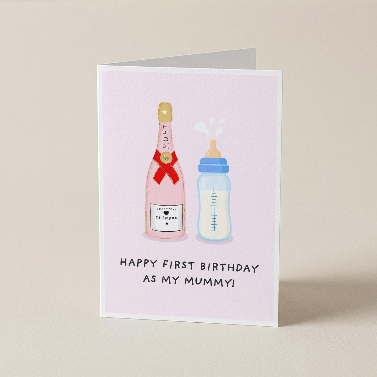 A folded greeting card standing upright on a light beige surface. The card has a pale pink background and features an illustration of a champagne bottle and a baby bottle with the text 'HAPPY FIRST BIRTHDAY AS MY MUMMY!'.