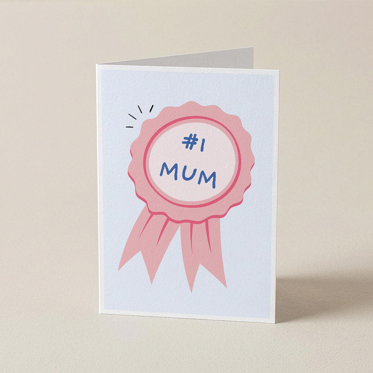A folded greeting card standing upright on a light beige surface. The card features a pink ribbon badge illustration with the text '#1 MUM' in blue.