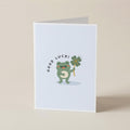 A folded white greeting card standing upright on a light beige surface. The front features a cute green cartoon frog holding a four-leaf clover with the words 'GOOD LUCK!' curved above it.