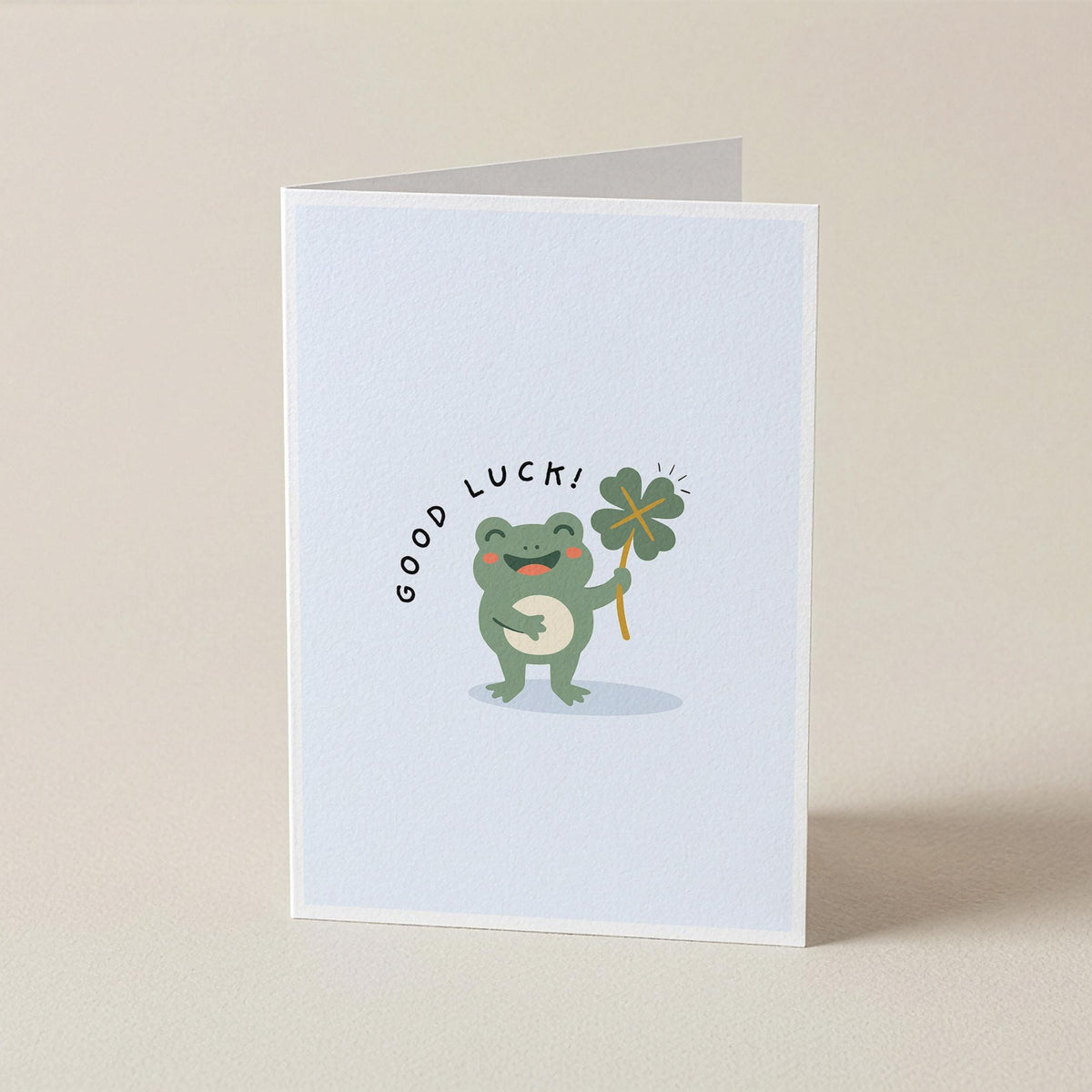 A folded white greeting card standing upright on a light beige surface. The front features a cute green cartoon frog holding a four-leaf clover with the words 'GOOD LUCK!' curved above it.