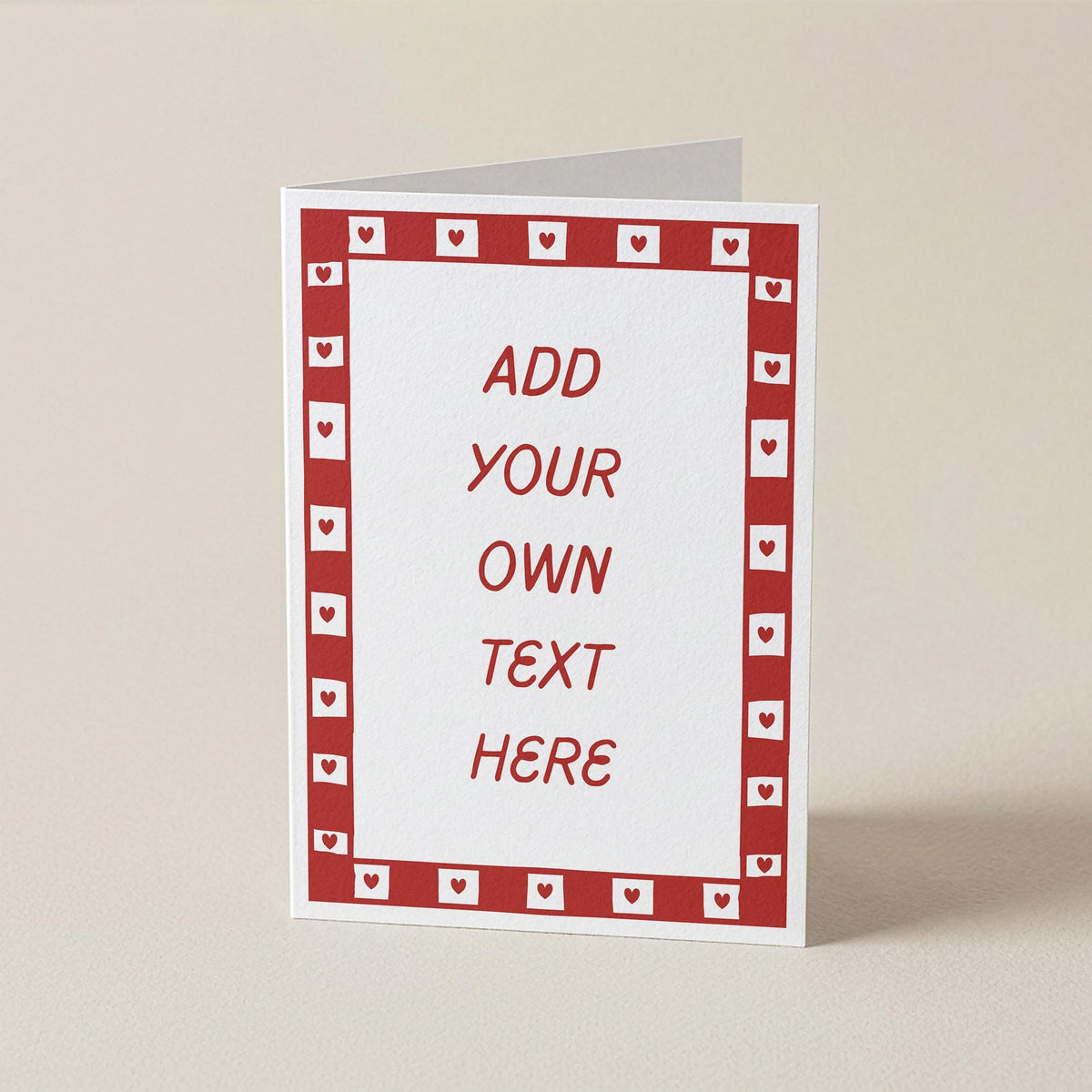 A customizable greeting card standing upright on a neutral beige background. The card has a white center with red placeholder text reading 'ADD YOUR OWN TEXT HERE' and a red border pattern of small hearts.