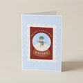 A folded Christmas greeting card standing upright on a light beige surface. The card has a pale blue background with subtle white snowflake dots, a central red rectangular panel, a scalloped white border, and an illustration of a snowman in a green hat and scarf inside a snow globe.