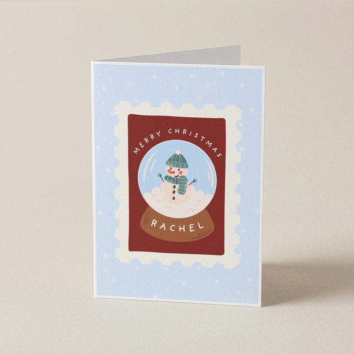 A folded Christmas greeting card standing upright on a light beige surface. The card has a pale blue background with subtle white snowflake dots, a central red rectangular panel, a scalloped white border, and an illustration of a snowman in a green hat and scarf inside a snow globe.