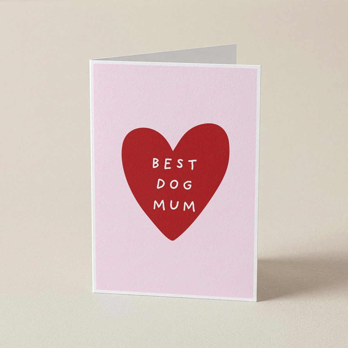 A folded greeting card standing upright on a light beige surface. The card has a pale pink background with a large red heart in the center containing the white text 'BEST DOG MUM'.